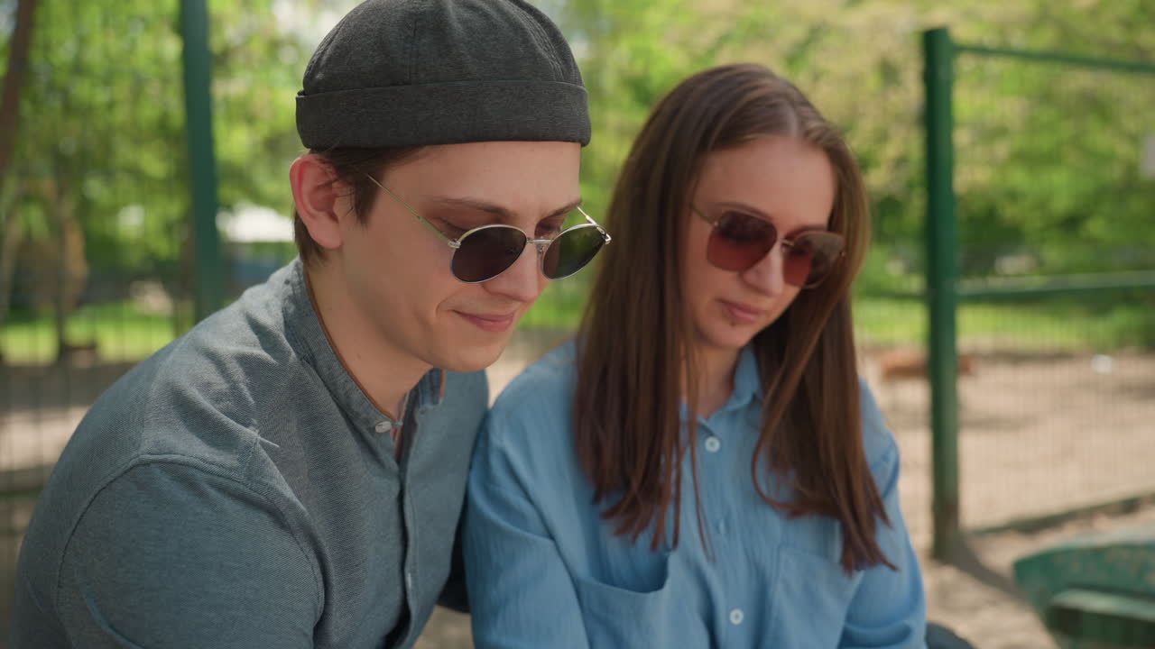 Joven pareja sentada en un banco planeando una ruta en el teléfono, ambiente relajado de parque veraniego, gafas de sol, ropa informal, luz suave del sol entre los árboles, conversación centrada y sonrisas delicadas.