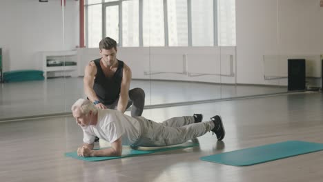 Un-Hombre-De-Edad-Avanzada-Entrenando-En-Plancha-Con-Un-Entrenador-Deportivo-En-El-Gimnasio-Juntos