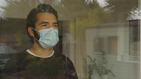 hombre mirando por la ventana con una máscara facial en cuarentena