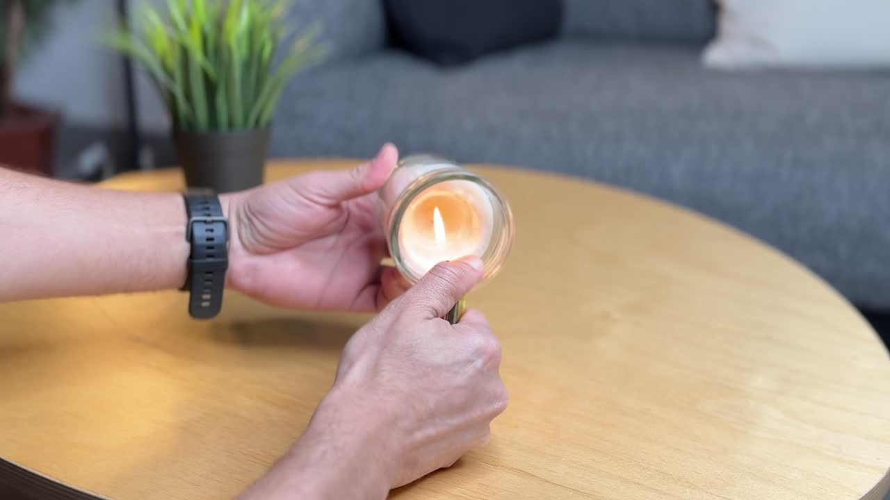 4K close-up of hands lighting a candle in a glass jar on a wooden table with a cozy background. Ideal for themes of relaxation, home life, rituals, or ambient lifestyle visuals.