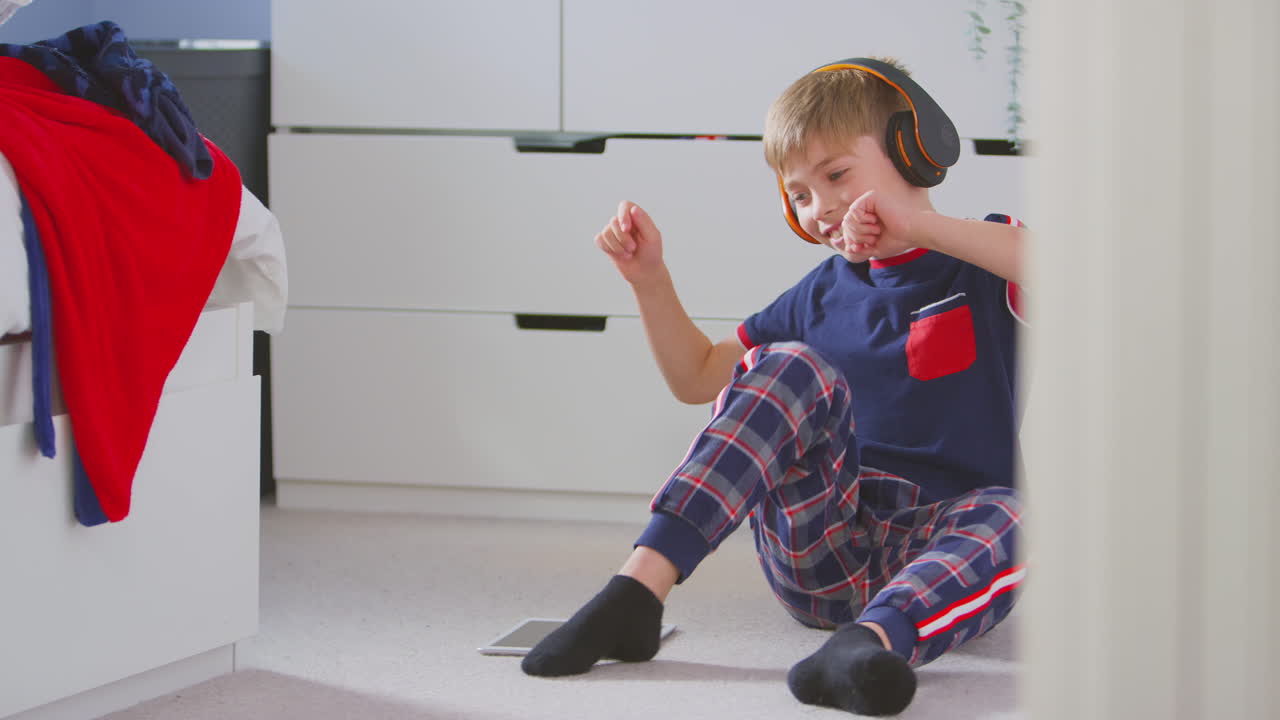 Boy At Home In Bedroom Wearing Wireless Headphones Listening To Music On Digital Tablet