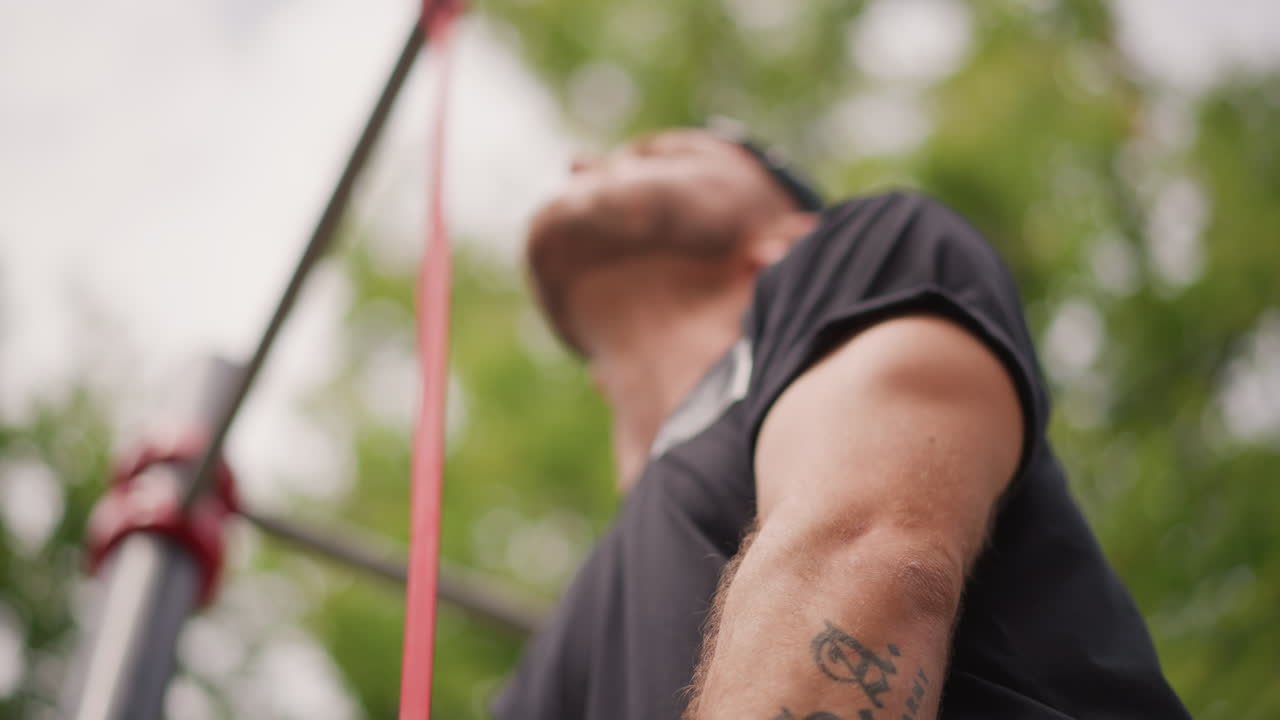 Outdoor Scene With Inked Man Adjusting Gear, Tattooed Male Athlete Finetunes Equipment Beneath Leafy Trees, Inkcovered Man Brightens His Training Setup Outdoors Amidst Lush Canopy Sunlight