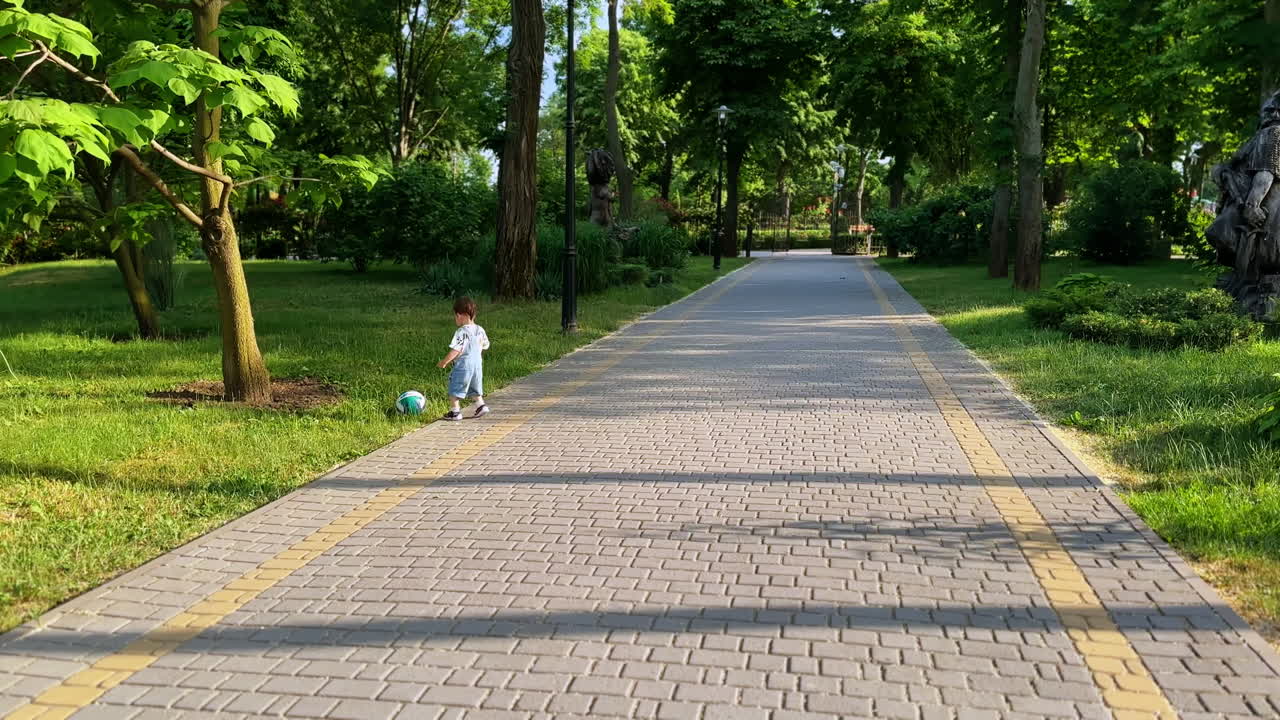 Little boy running funnily by the park. Kid in jeans romper picks up the ball from grass and runs up to camera.