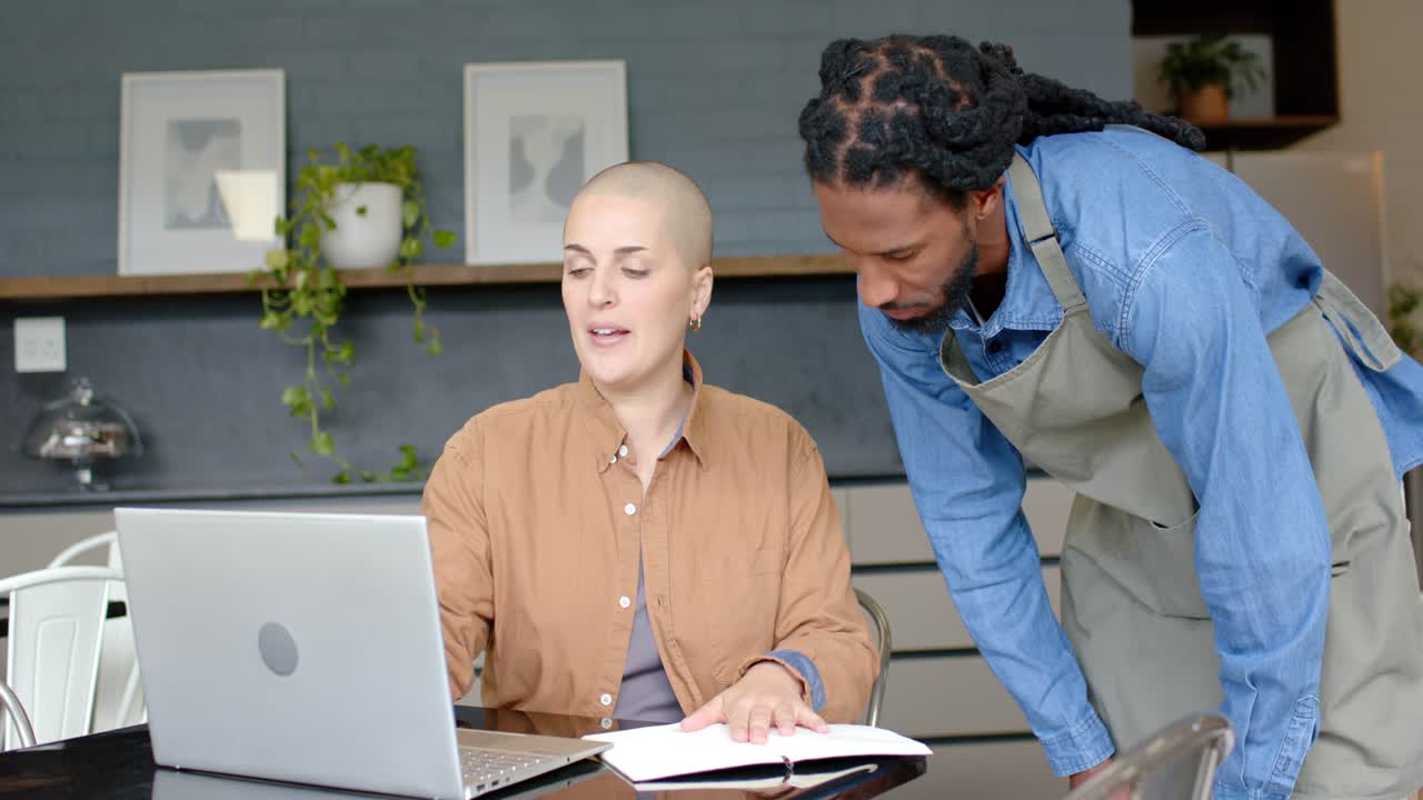 Diverse coworkers planning project with man approaching, writing in notebook and navigating laptop