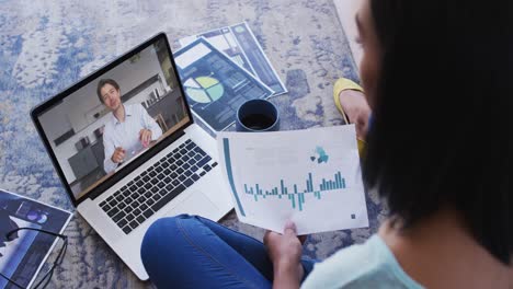 Mixed-race-businesswoman-sitting-on-floor-using-laptop-having-video-call-with-male-colleague