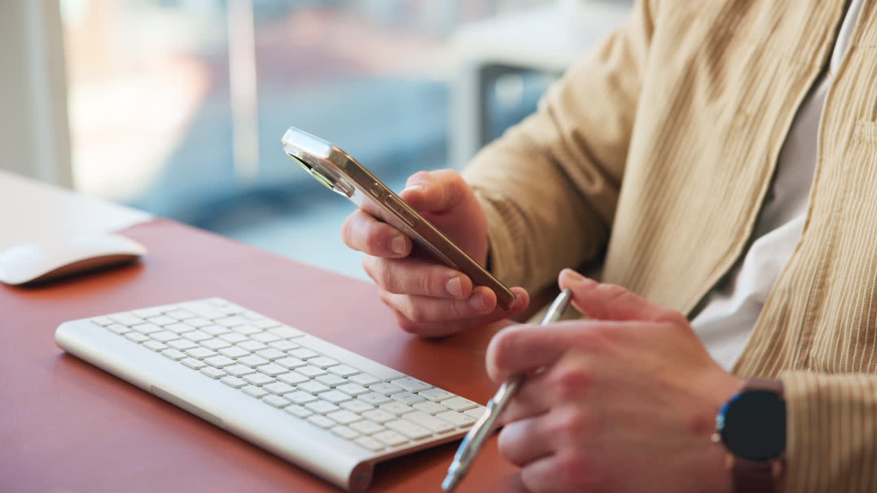 Close-up of Person's Hands Holding Smartphone and Pen at Desk
