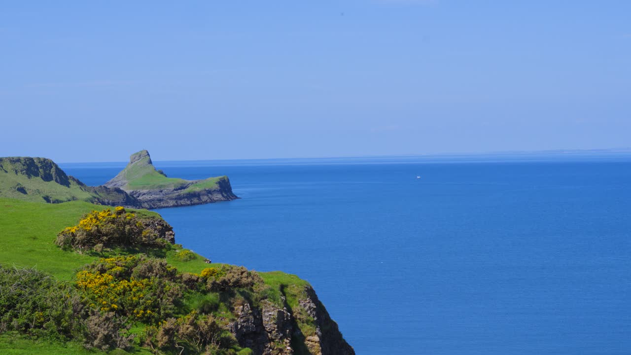 Slow Motion Pan Across Rhossili Bay Revealing Worm's Head Landmark with Brambles on Cliff Tops and Clear Bue Sky and Sea.