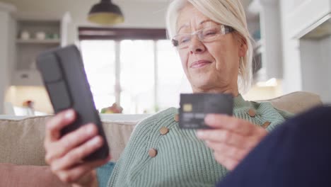 happy senior caucasian woman on sofa in living room, using smartphone and holding credit card