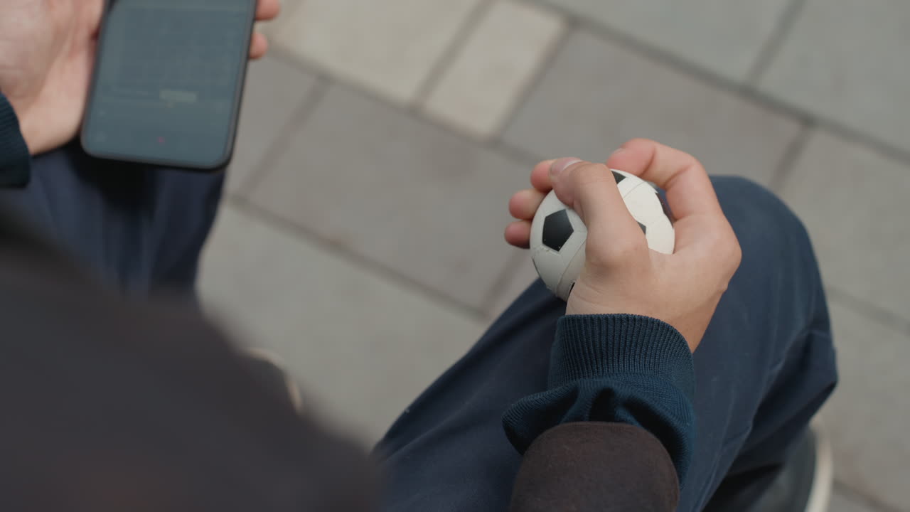 primo piano delle mani di un uomo bianco che tiene in mano un mini pallone da calcio e uno smartphone, le dita stringono e ruotano il pallone per migliorare la manualità, enfasi sulla texture e il movimento, calma terapeutica, morbido effetto bokeh su una panchina urbana