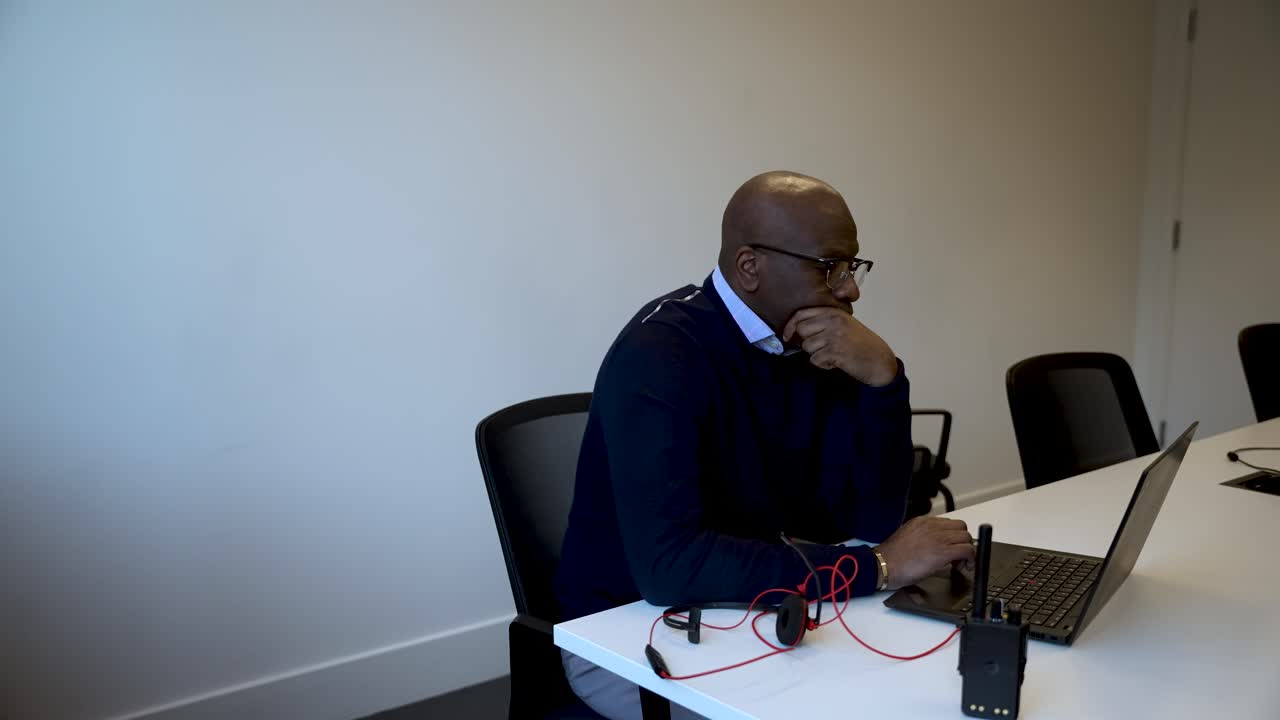 A man is seated at a desk in a modern office space, focused on his laptop, with a pair of red earphones and a walkie-talkie placed next to him on the desk