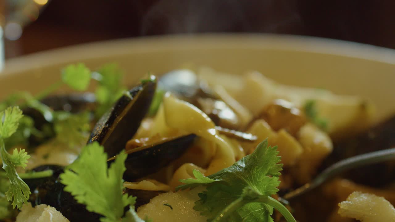 A close-up view of a hand using a spoon to serve hot seafood chowder with mussels, fish, squid, and fresh cilantro in warm, natural lighting