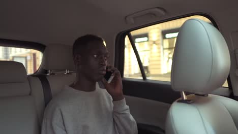 Calm-handsome-african-man-in-casual-clothes-talking-on-cellphone-while-riding-in-backseat-of-taxi-cab.-Cheerful-black-passenger-guy-answering-call-phone-during-road-trip-in-the-city