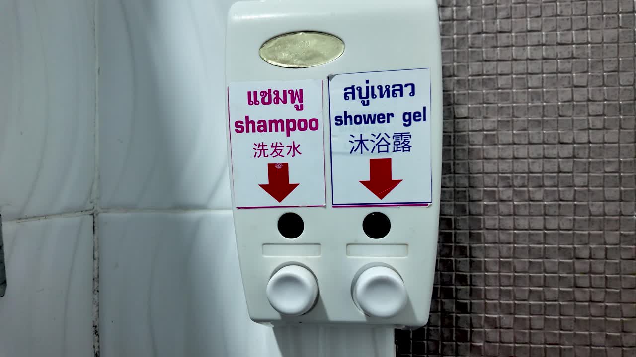 A white plastic soap dispenser with shampoo and shower gel is mounted on the wall in a Thai hotel bathroom. This hygiene product is commonly found in travel and tourism accommodations