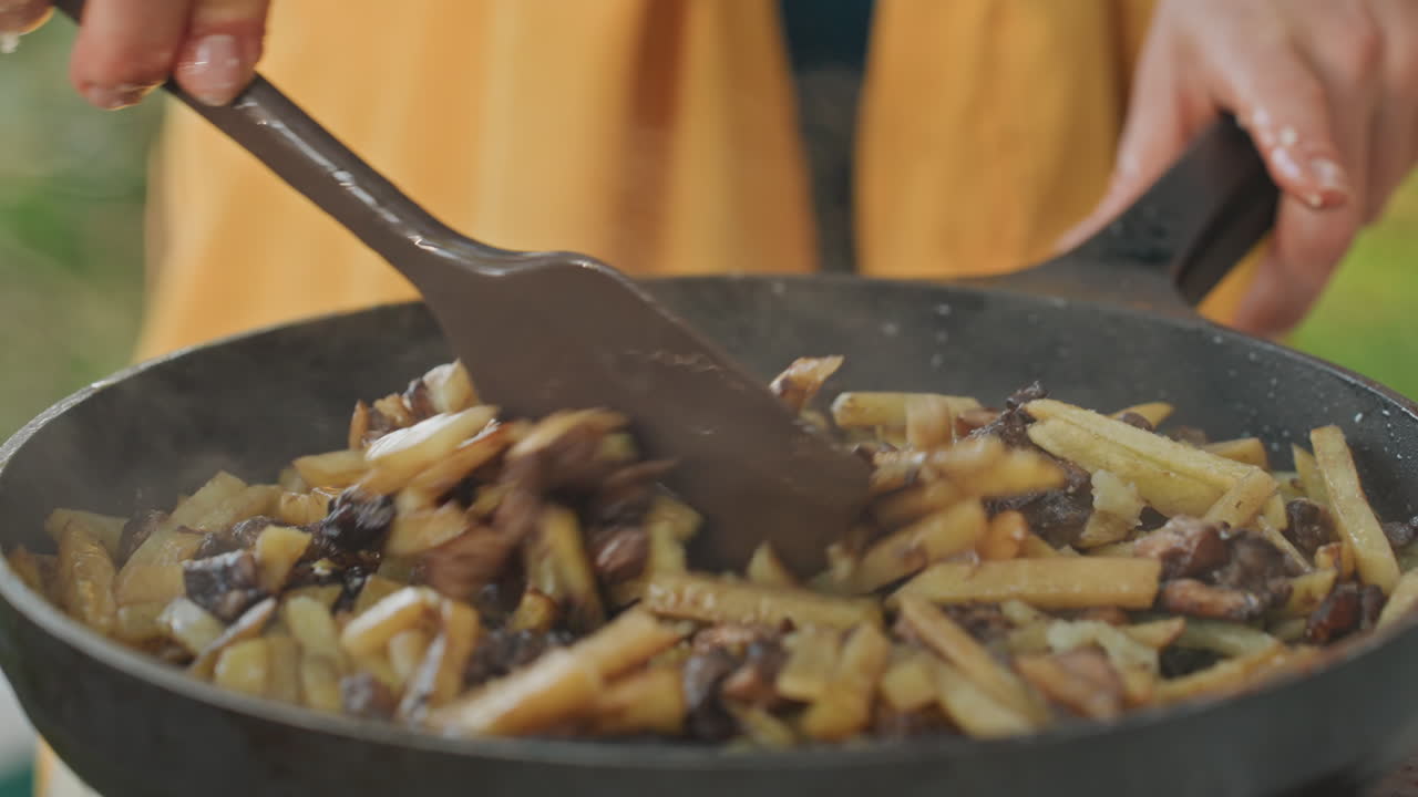 Sizzling fries and mushrooms in pan, hands stir with wooden spatula in castiron skillet, steam rising over golden crisp fries and sauteed mushrooms, smoky aroma fills campsite, cozy rustic camping
