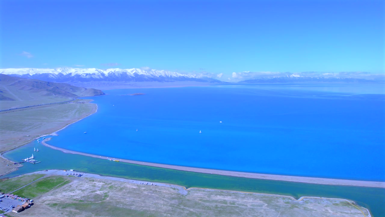 Flygfoto över Sayram-sjön i Yili, Xinjiang, med Tianshan-snöberg. En yacht seglar på den alpina sjön som är omgiven av barrskog och gräsmark. Nordvästra Kinas naturliga landskap vid Sayram-sjön.