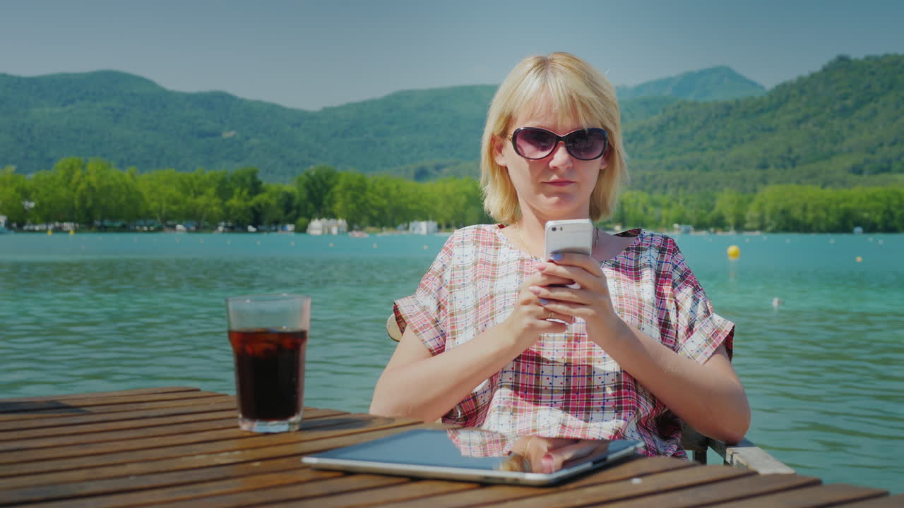 Ung kvindelig turist slapper af et smukt sted ved søen og bjergene. Hun bruger telefonen, siddende.