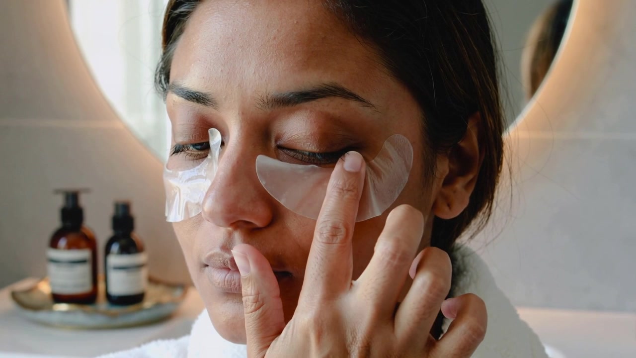 Raising right hand, woman pressing under-eye patches at vanity ensuring adhesion