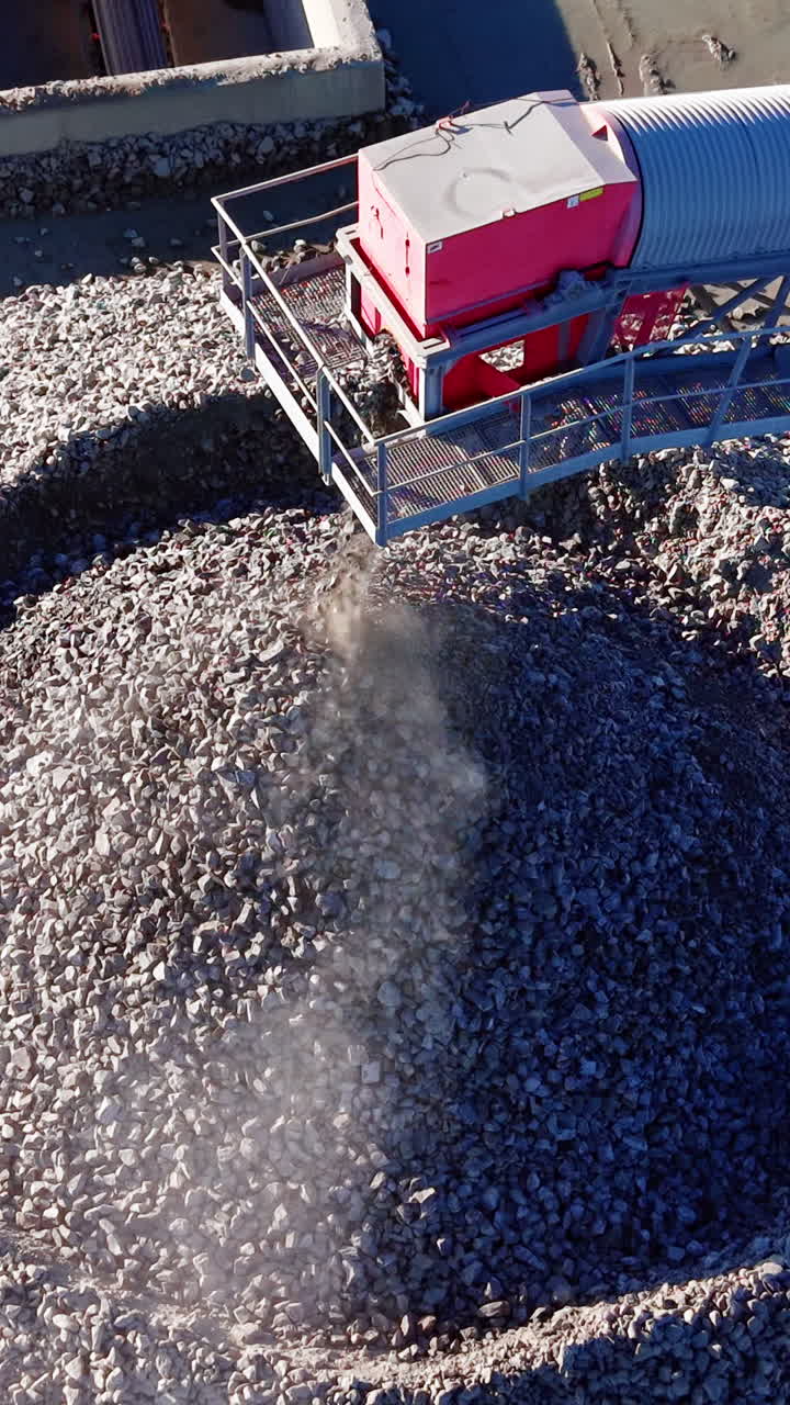 Testa di nastro trasportatore rosso che rilascia roccia minerale frantumata, creando polvere e un grande cumulo di aggregati. Primo piano del nastro trasportatore che scarica rocce su un mucchio. Video verticale.
