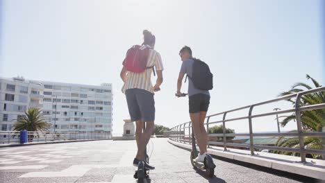 Happy-diverse-gay-male-couple-using-scooters-at-promenade-by-the-sea,-slow-motion