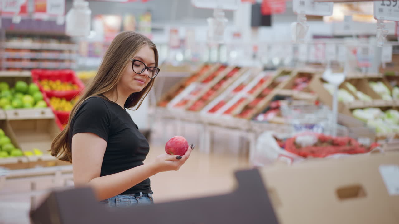 trabajador remoto sonríe mientras recoge la manzana roja de la exhibición, observándola y dejándola caer, etiqueta de precio colgada en el fondo con señal visible en el entorno de la tienda brillante