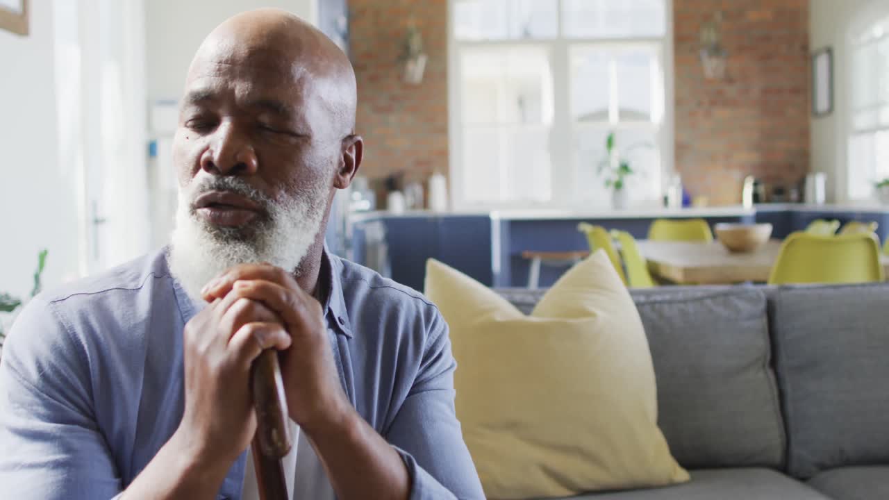 Thoughtful senior african american man in living room sitting on sofa, holding walking cane