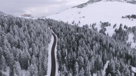 Aerial-revealing-shot-on-a-mountain-road-surrounded-by-snowy-forest-ending-on-a-mountain-hut-during-winter