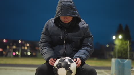 un hombre se sienta con la cabeza hacia abajo, sosteniendo una pelota de fútbol desgastada entre sus piernas, el fondo presenta una vista borrosa de un estadio y un poste de gol