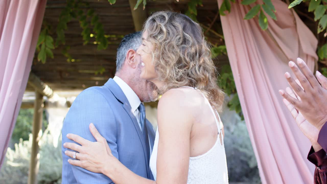Coppia che si abbraccia, sciogliendosi dopo l'applauso degli invitati, girandosi, sorridendo e tenendosi per mano sotto un pergolato