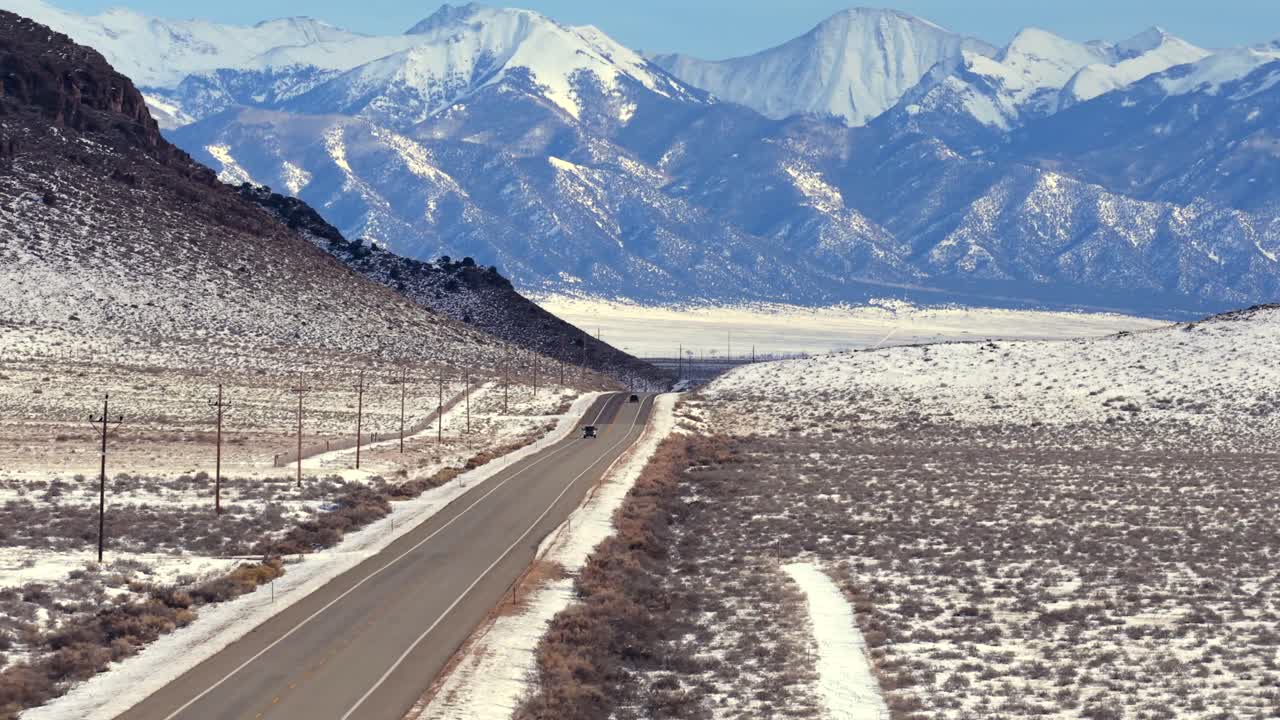 A lone truck drives down a remote highway as the majestic, monolithic snow-capped Crestone Group mountains rise dramatically. Captures the vast winter scale of the Sangre de Cristo Range, Colorado