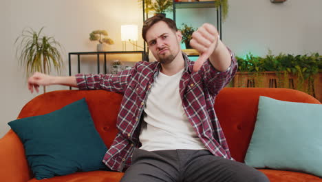 a man sitting on a couch, giving a thumbs down gesture. he looks tired and frustrated.