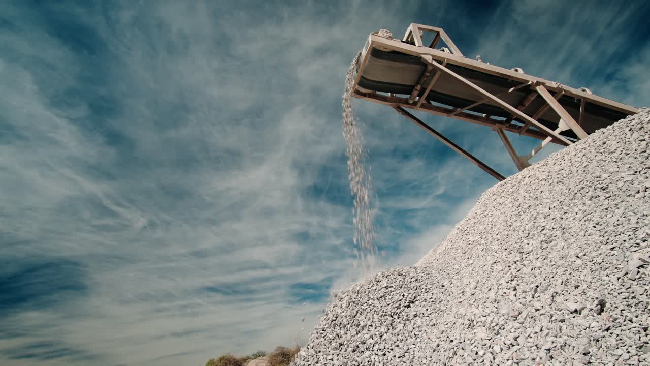 conveyor belt of a rock crusher dumping aggregate in slow motion onto a pile under a blue sky with clouds