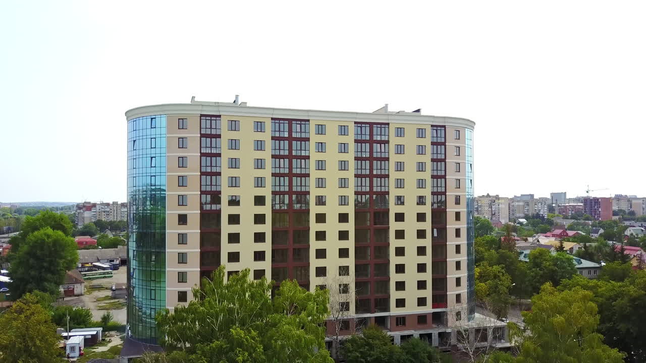 New modern city architecture. Multi-storied residential building with rounded sides. Lush greenery surrounding the house. Cityscape at backdrop.