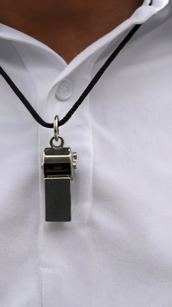 Video vertical: Colgante de silbato balanceándose en un cordón negro a la altura del pecho, con una camisa blanca moviéndose ligeramente.