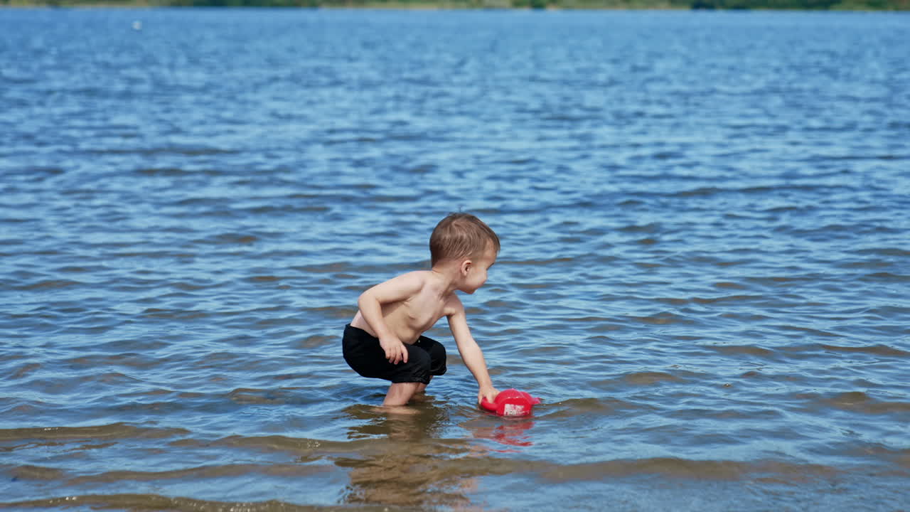 Smiling happy toddler stands in river to his ankles. Playful kid throws his watering can into water and watches it float.