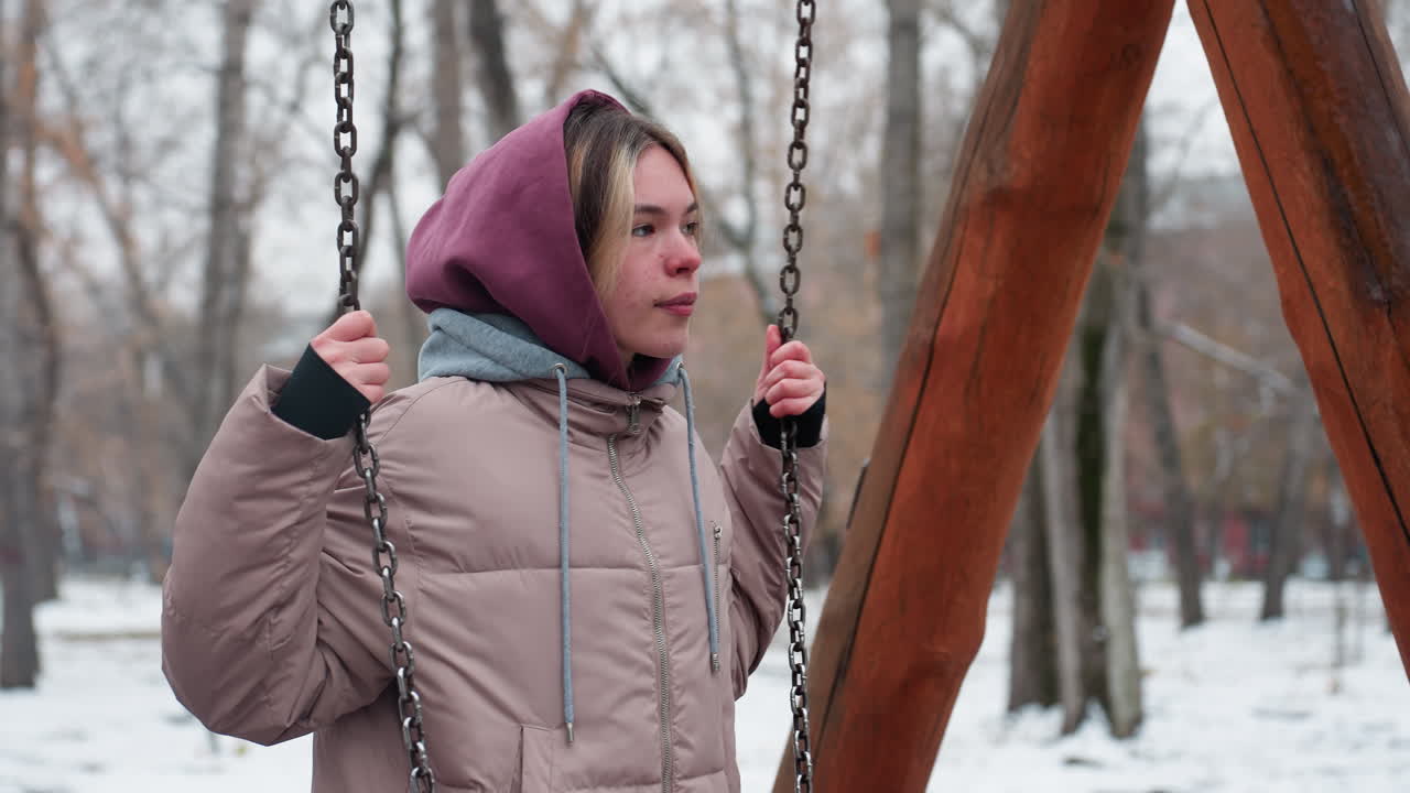 молодая женщина в зимнем наряде держит летучие цепи, кладя колено на железную люльку в городском парке, покрытой снегом земле, голых деревьях и жилых зданиях