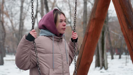 joven con traje de invierno sostiene cadenas de columpio mientras coloca su rodilla en el columpio de hierro en el parque urbano, el suelo cubierto de nieve, los árboles desnudos y los edificios residenciales