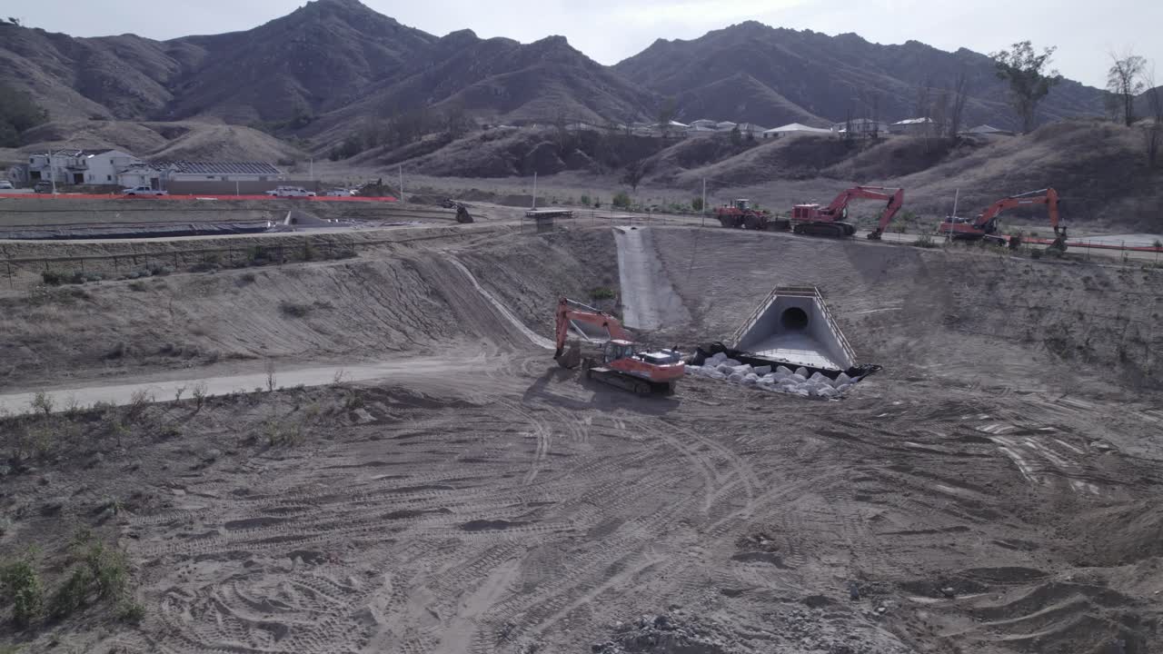 A drone circles around an excavator parked within a sizable pit adjacent to a water runoff drain