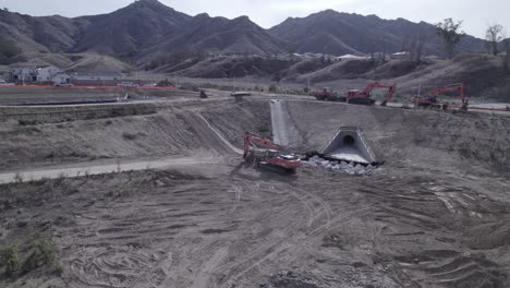 a drone circles around an excavator parked within a sizable pit adjacent to a water runoff drain