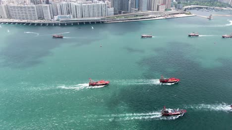 Convoy-of-local-Fishing-boats-causing-in-Hong-Kong-Victoria-bay,-with-city-skyline-in-the-horizon,-Aerial-view