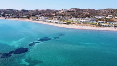 cinematic drone shot of paradisiacal greek beach with a beautiful sea and some houses on the shore