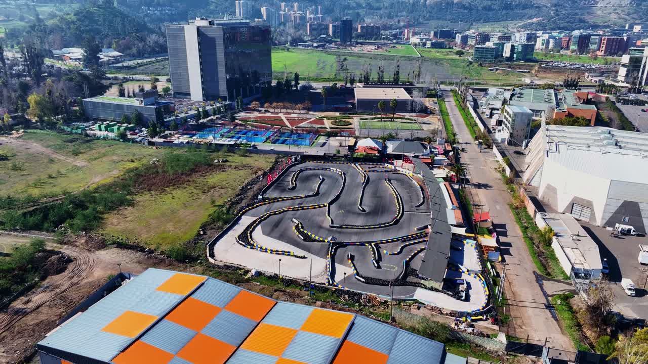 Orbiting drone aerial of outdoor go-kart racing circuit with curved track layout, barriers, and surrounding urban recreational facilities