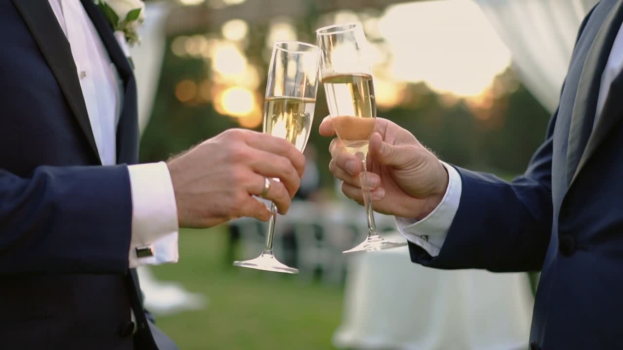 Toasting, two men in tuxedos raising flutes and clinking at reception lawn, boutonniere visible