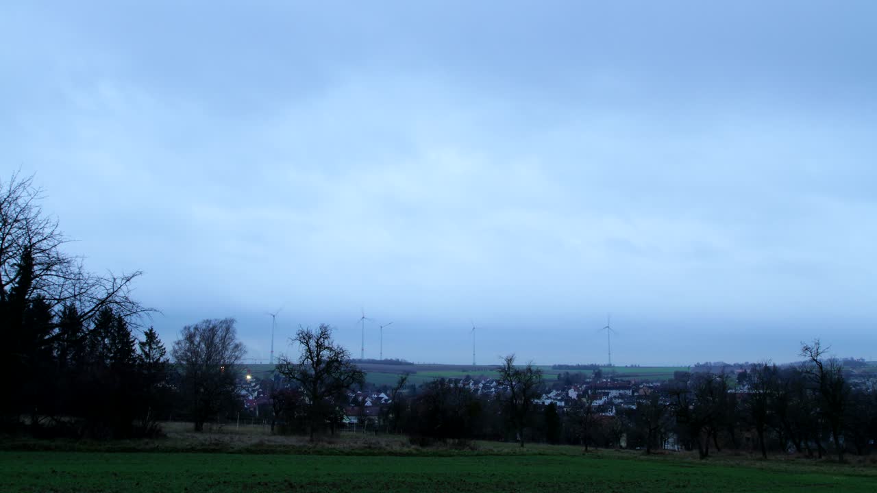 Timelapse of an German landscape with a small town as clouds move past and it gets dark with wind turbines in the background.