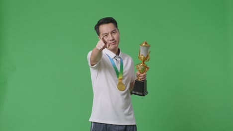 hombre asiático con una medalla de oro y trofeo desaprobando sin señal del dedo índice y sonriendo a la cámara en el fondo de la pantalla verde en el estudio