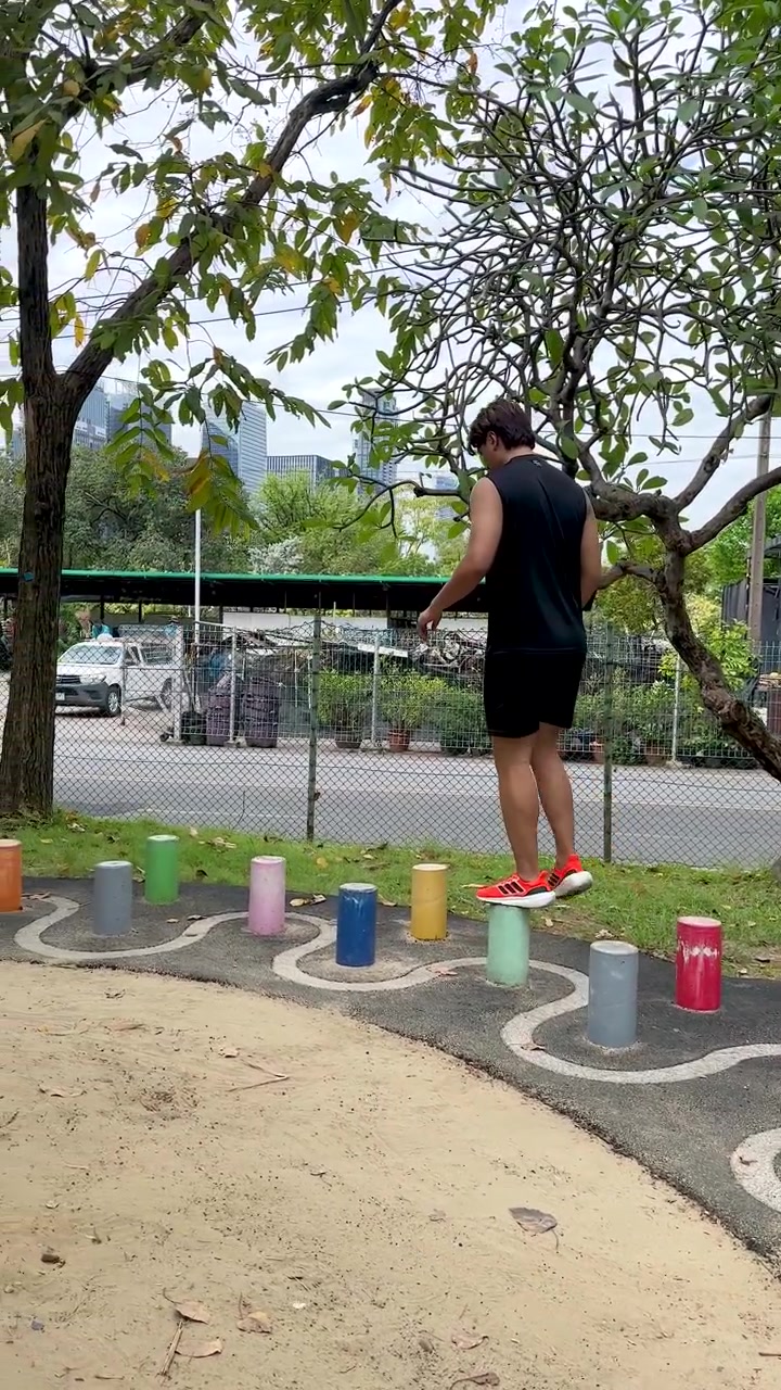 Um homem asiático em roupa esportiva preta se equilibra em postes coloridos durante exercícios em parque ao ar livre.