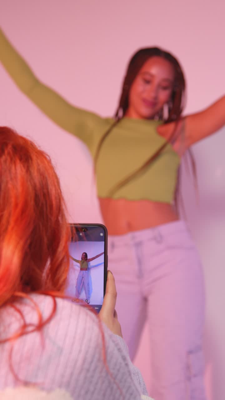 fotografía de estudio de video vertical de una mujer tomando una foto de una amiga bailando en un teléfono móvil contra un fondo rosa