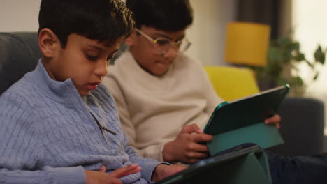 two young boys sitting on sofa at home playing games or streaming onto digital tablets 16