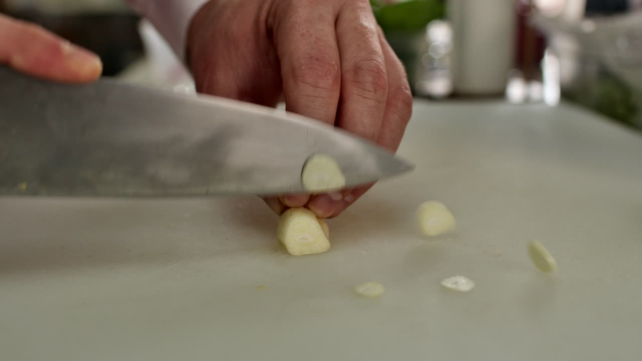 close-up of a chef's hands finely slicing fresh garlic cloves on a white cutting board in a generic kitchen. Essential culinary prep for a gourmet tomato sauce recipe