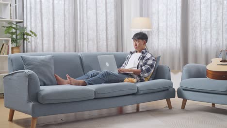 full body of asian teen boy with snacks typing on a laptop while lying on sofa in the living room at home