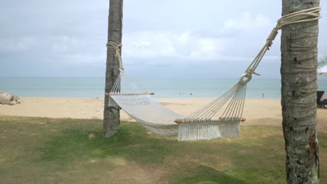 Leere-Hängematte-Auf-Grünem-Rasen-In-Der-Nähe-Von-Sandigem-Tropischem-Strand,-Schwingendes-Bett-Durch-Blaues-Meer-Auf-Exotischen-Reisen-Sommerzielferien